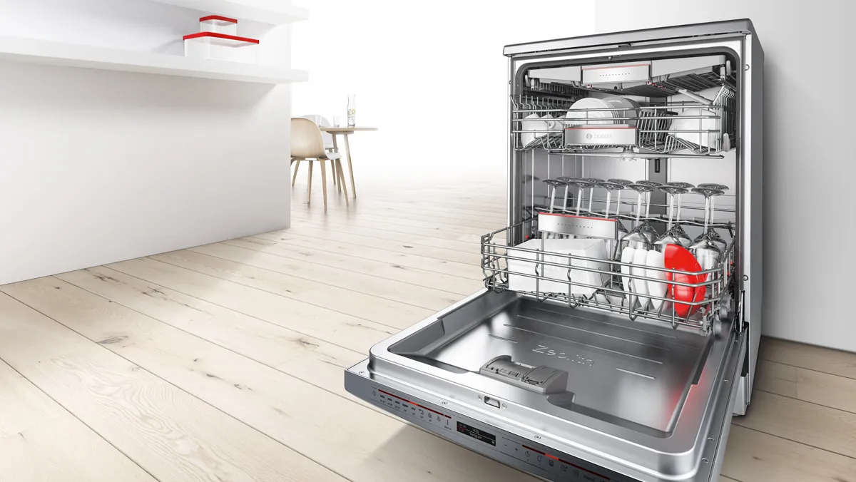 Freestanding stainless steel Bosch dishwasher with its door open, loaded with dishes on a light wooden floor.