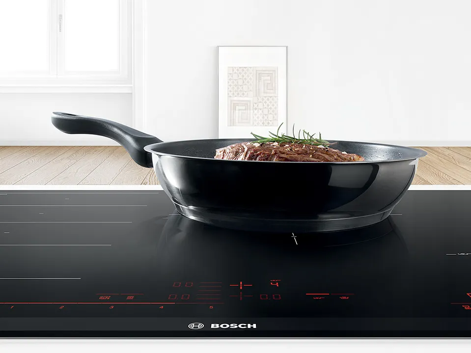 A black frying pan with a piece of meat and herbs sits on a modern Bosch induction cooktop in a bright kitchen.