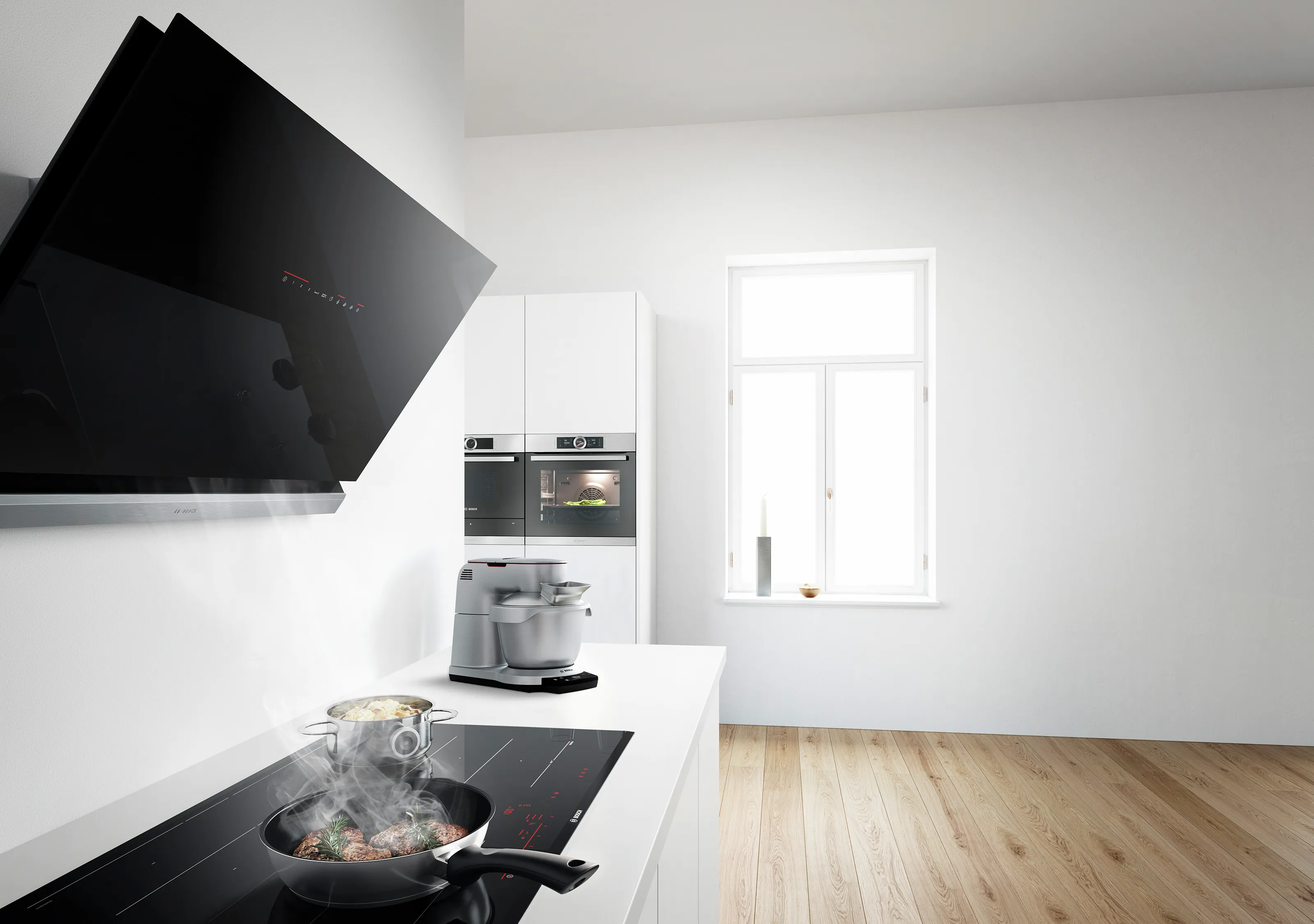 A wide shot of a modern, bright white kitchen with light wood floors. In the foreground, food is sizzling in a pan on a black induction hob, with steam rising towards a sleek, black angled cooker hood. A silver Bosch food mixer sits on the white countertop. In the background, two built-in Bosch ovens are integrated into white cabinetry, and a bright window illuminates the room.
