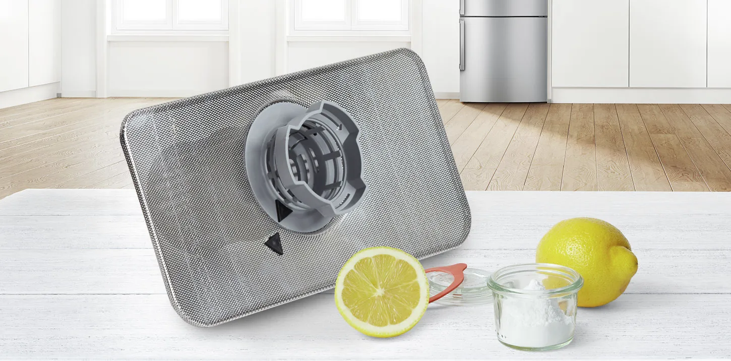 Dishwasher filter with lemon and baking soda on a kitchen table, modern fridge in background.