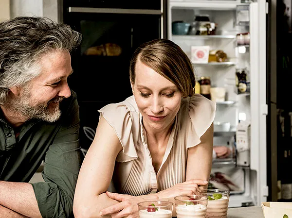 Two adults lean on a kitchen counter, smiling and looking at three small dessert jars in front of them, with an open, stocked refrigerator visible in the background.