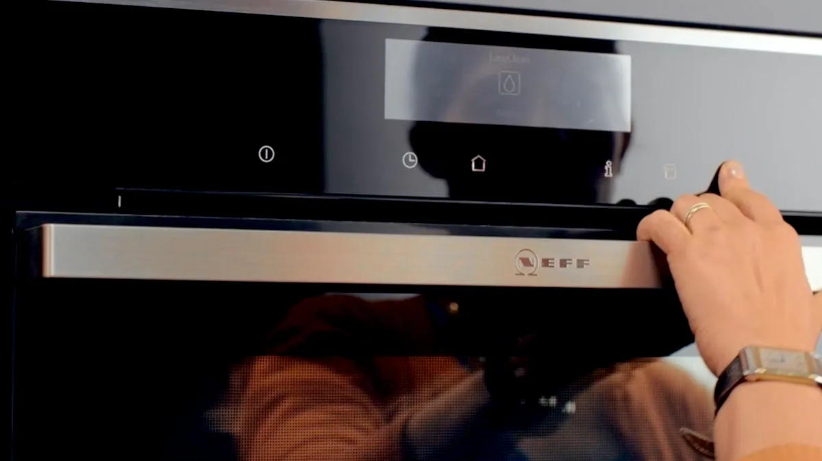 Close-up of a person’s hand turning a control dial on a black NEFF built-in oven, showing the stainless-steel handle and digital display.