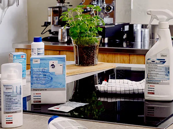Cleaning products around a black glass cooktop, with a potted herb and an espresso machine in the background.