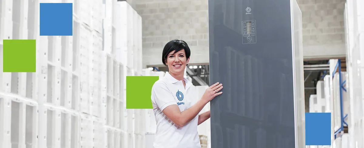 Trabalhador com um polo branco a segurar um eletrodoméstico alto e escuro, dentro de um armazém ou arrecadação com prateleiras brancas e caixas embaladas.