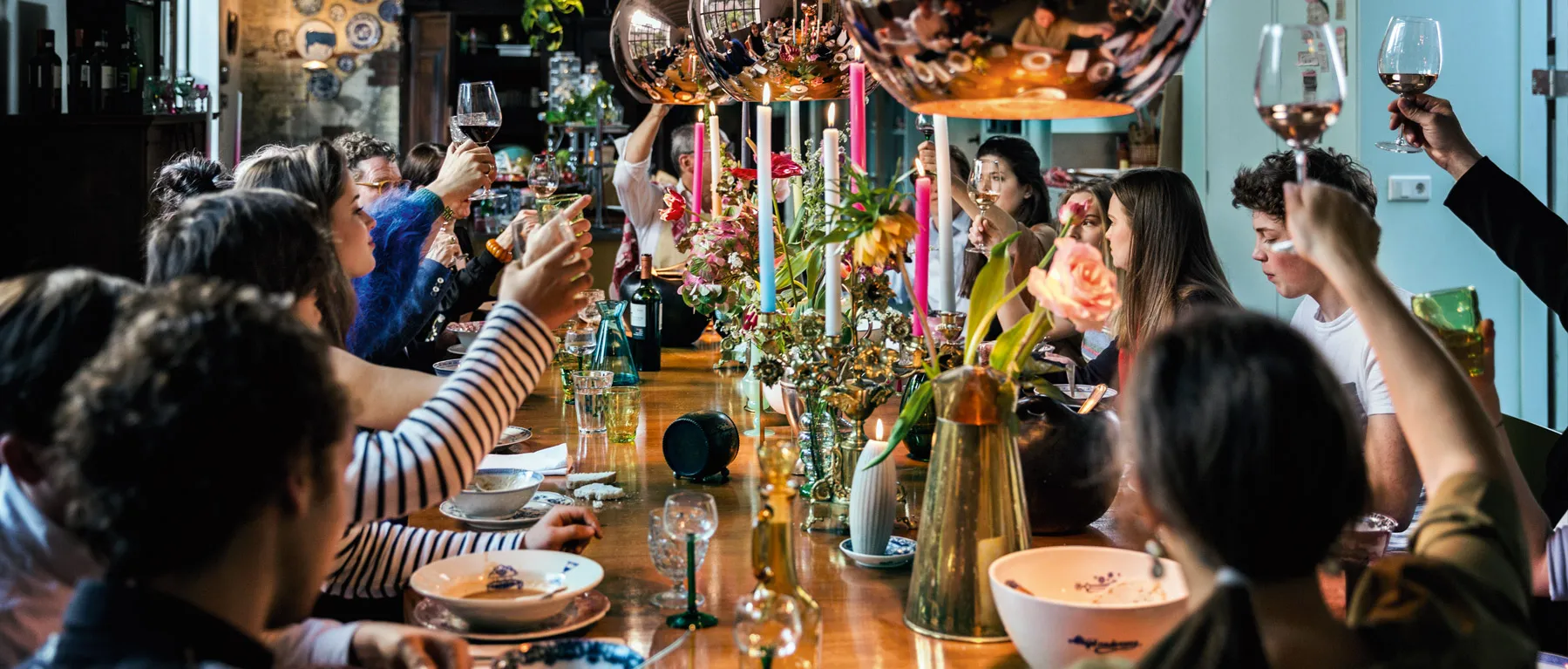 Un gruppo di persone che alzano i calici per un brindisi attorno a un lungo tavolo da pranzo decorato con fiori e candele.