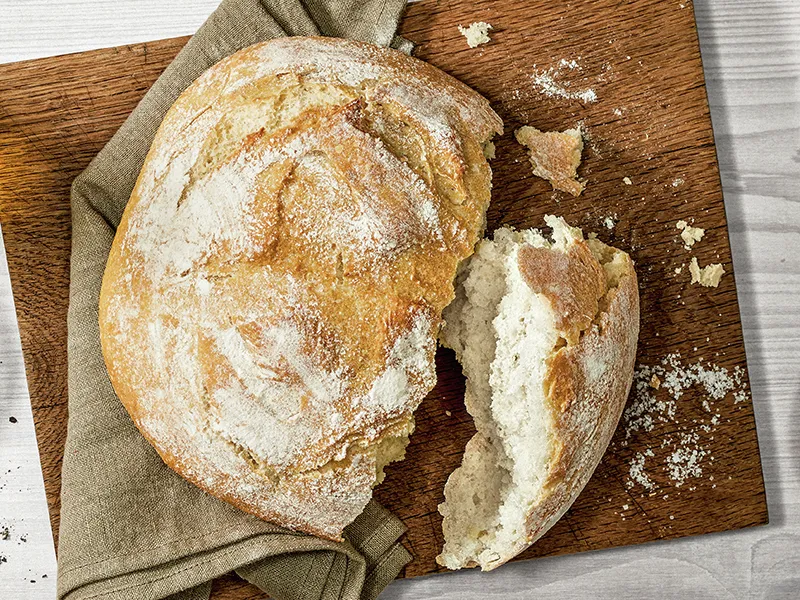 Pane artigianale appena cotto su tavola di legno, con una fetta di taglio che rivela l'interno morbido e arioso.