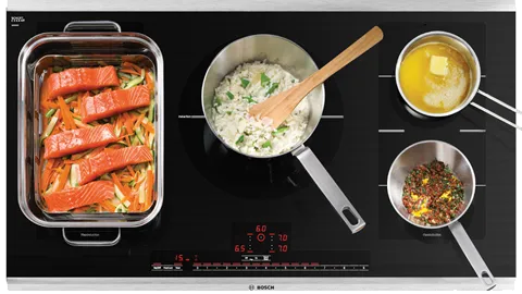 Vue de dessus d’une plaque de cuisson à induction avec du saumon dans un plat en verre, une casserole de riz crémeux aux fines herbes, un bol de beurre et une poêle contenant des épices.