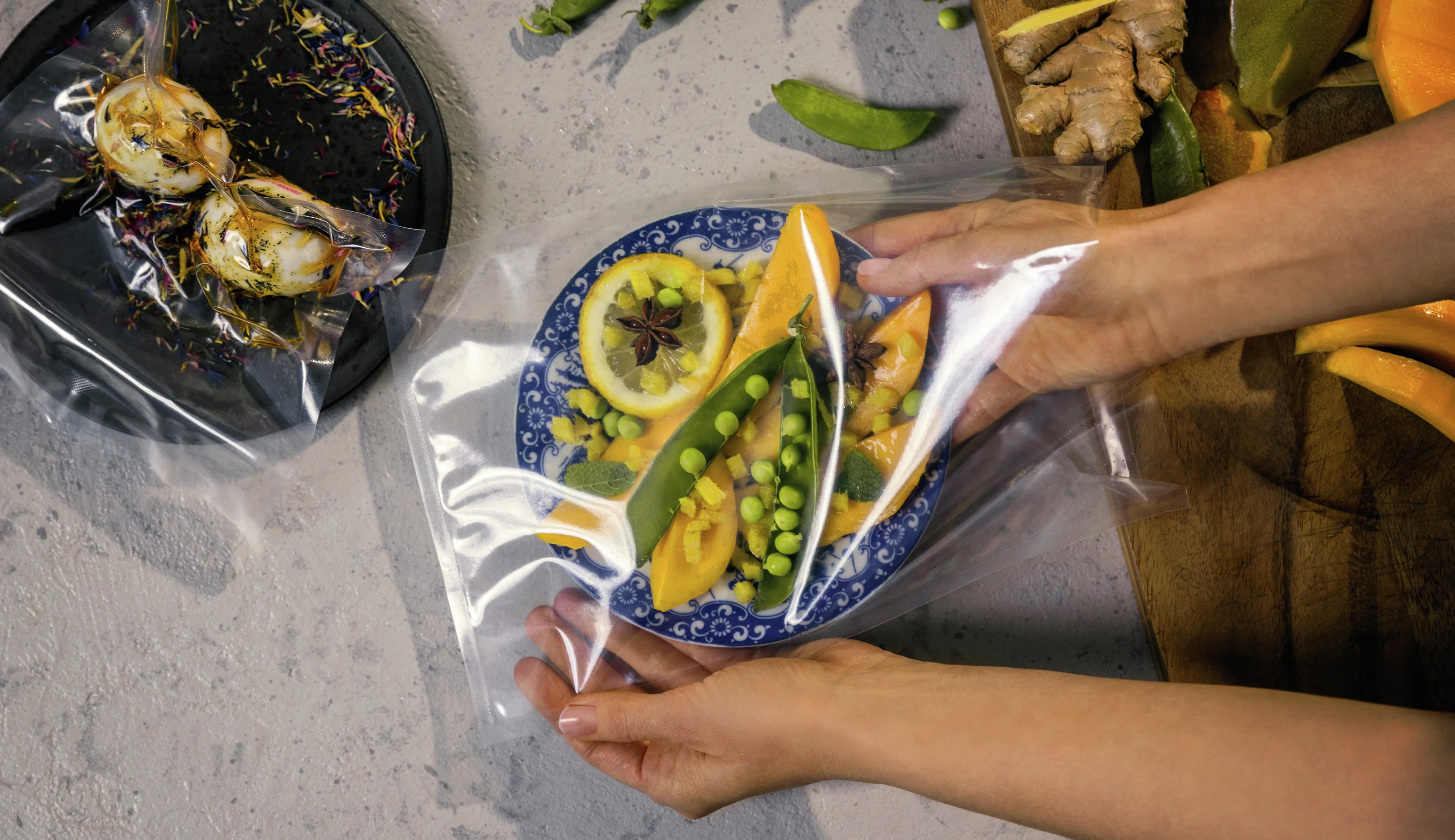 Una persona sigilla un piatto di frutta e verdura colorate in un sacchetto trasparente, circondato da ingredienti freschi su un piano di lavoro.