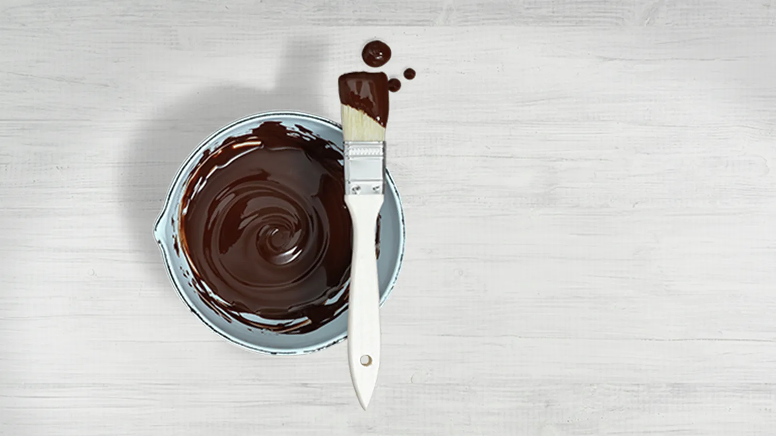 Bowl of melted chocolate with a brush on a light wooden table, chocolate drips beside.