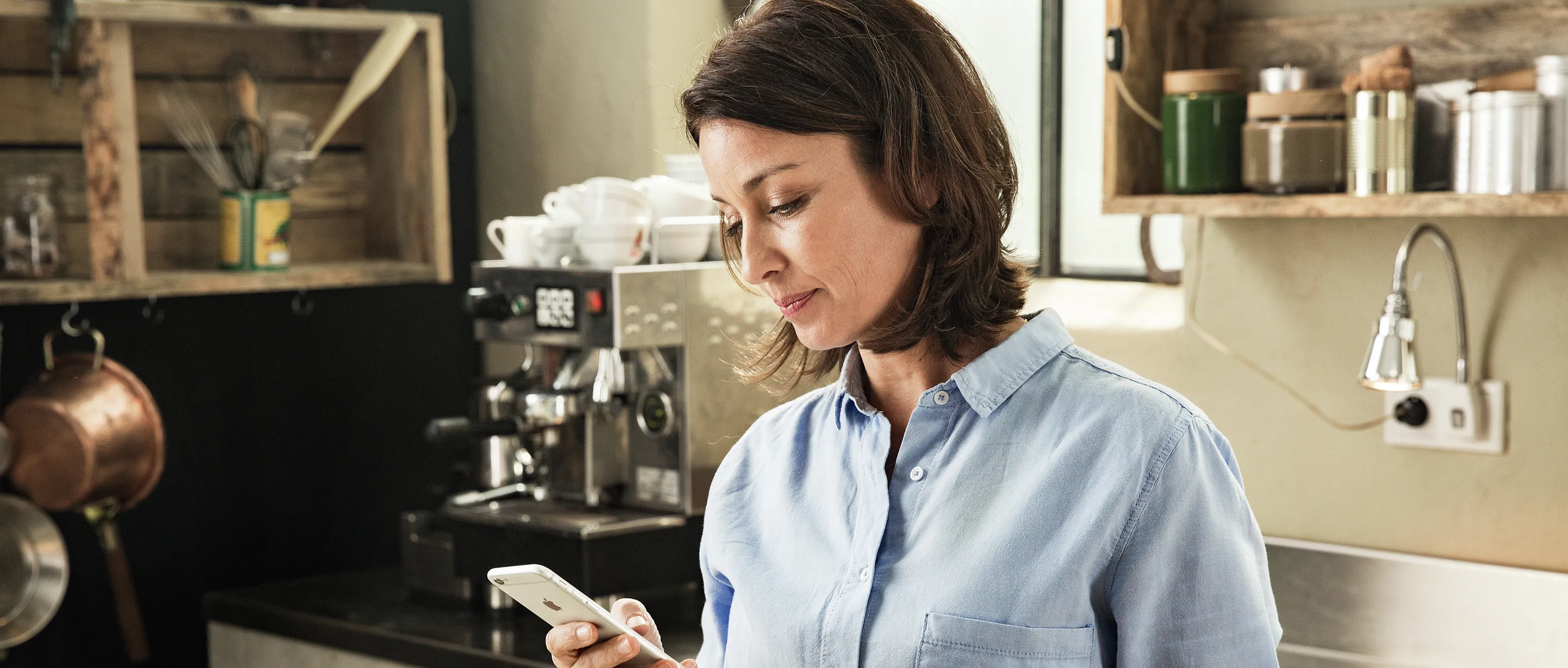 Ženska v kuhinji z vidnim pametnim telefonom, policami in napravami.