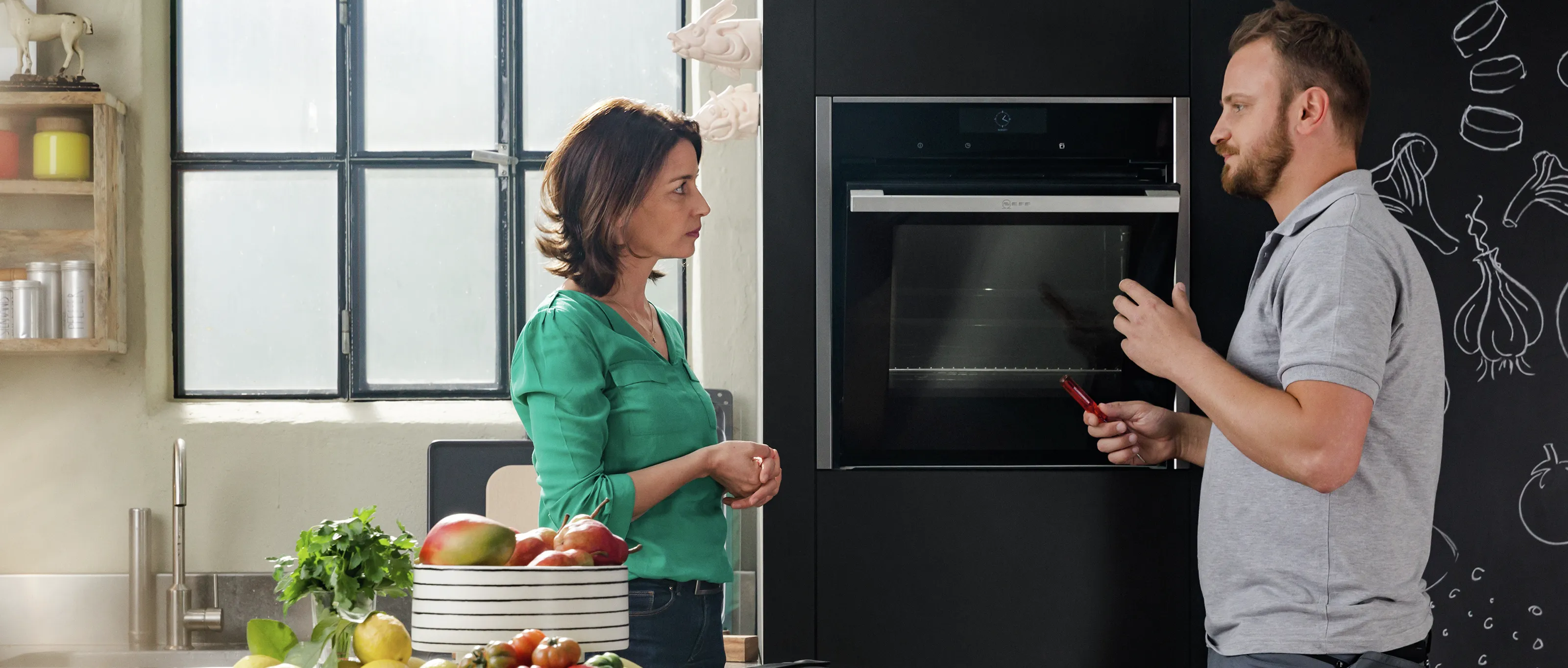 Couple dans la cuisine, femme tenant des légumes, homme ouvrant le four, appareils modernes, mur de craie.