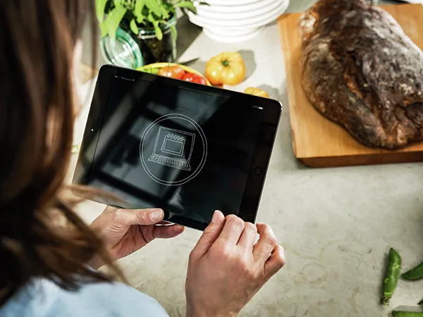 Hände halten eine Tablette mit einer Koch-App, umgeben von Kücheninhaltsstoffen und Utensilien.