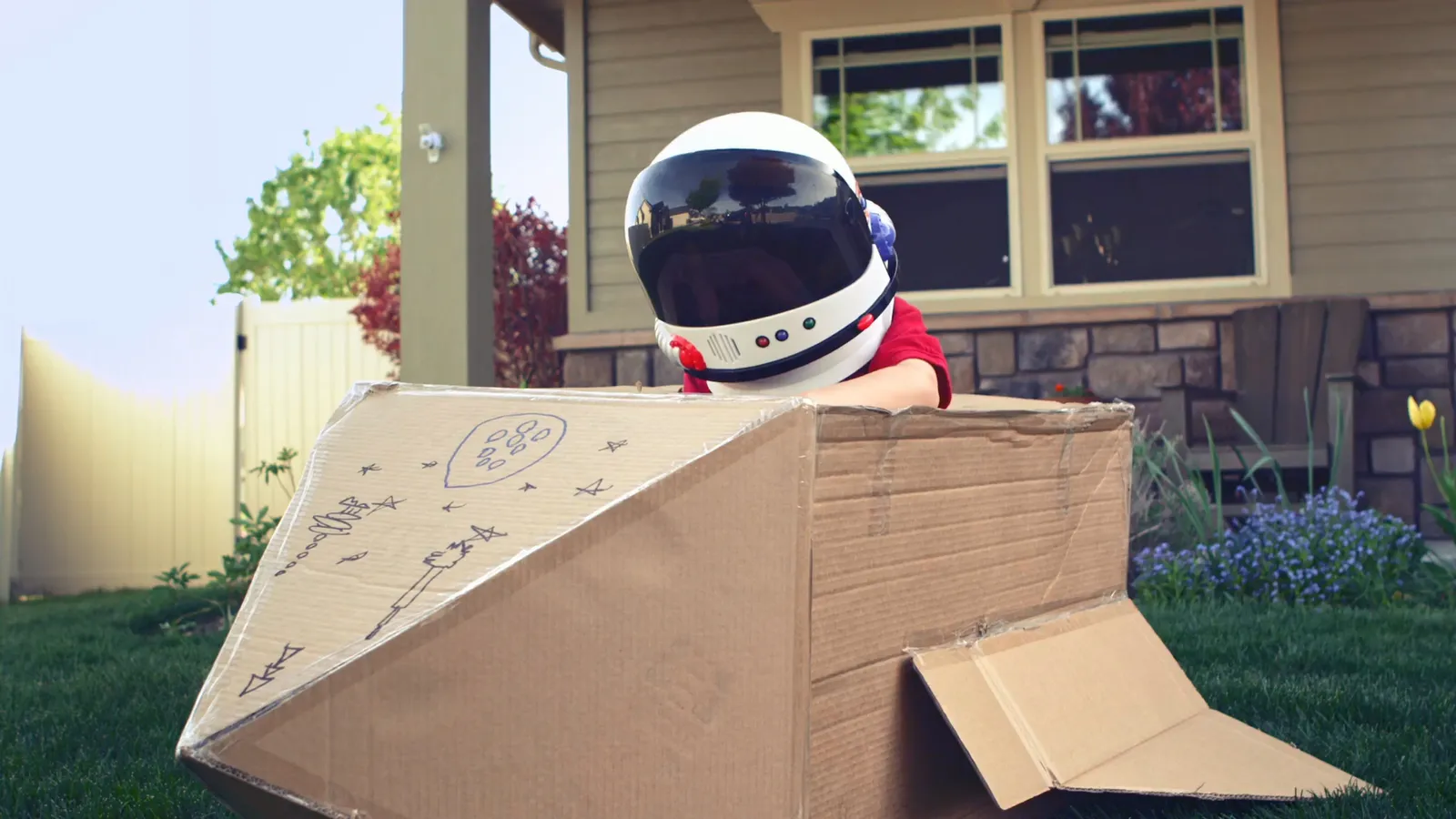 Bambino con un casco da astronauta che gioca in un'astronave di cartone nel giardino di casa.