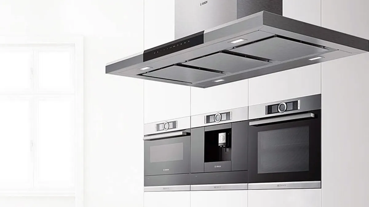 Moderne weiße Einbauküche mit einer hängenden Bosch Inselhaube aus Edelstahl im Vordergrund und einer Schrankwand mit integriertem Bosch Backofen, Kaffeevollautomaten und Mikrowelle.