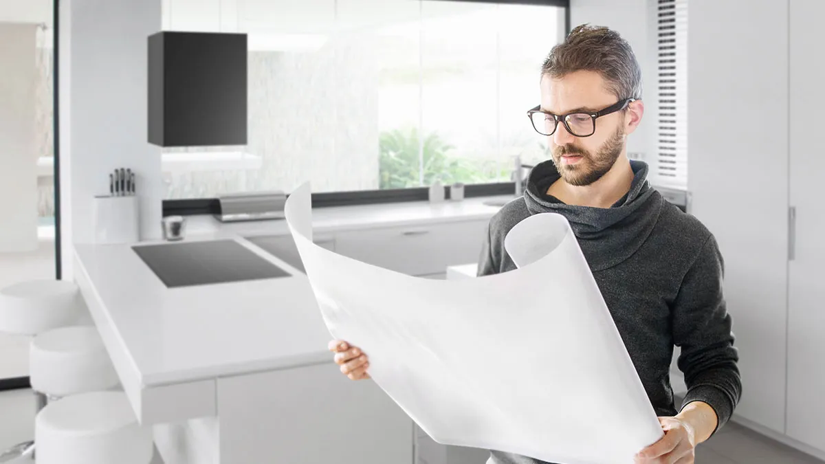 Ein Mann mit Brille prüft einen großen Bauplan im Vordergrund einer modernen, weißen Einbauküche mit Kochinsel und integriertem Bosch Kochfeld.