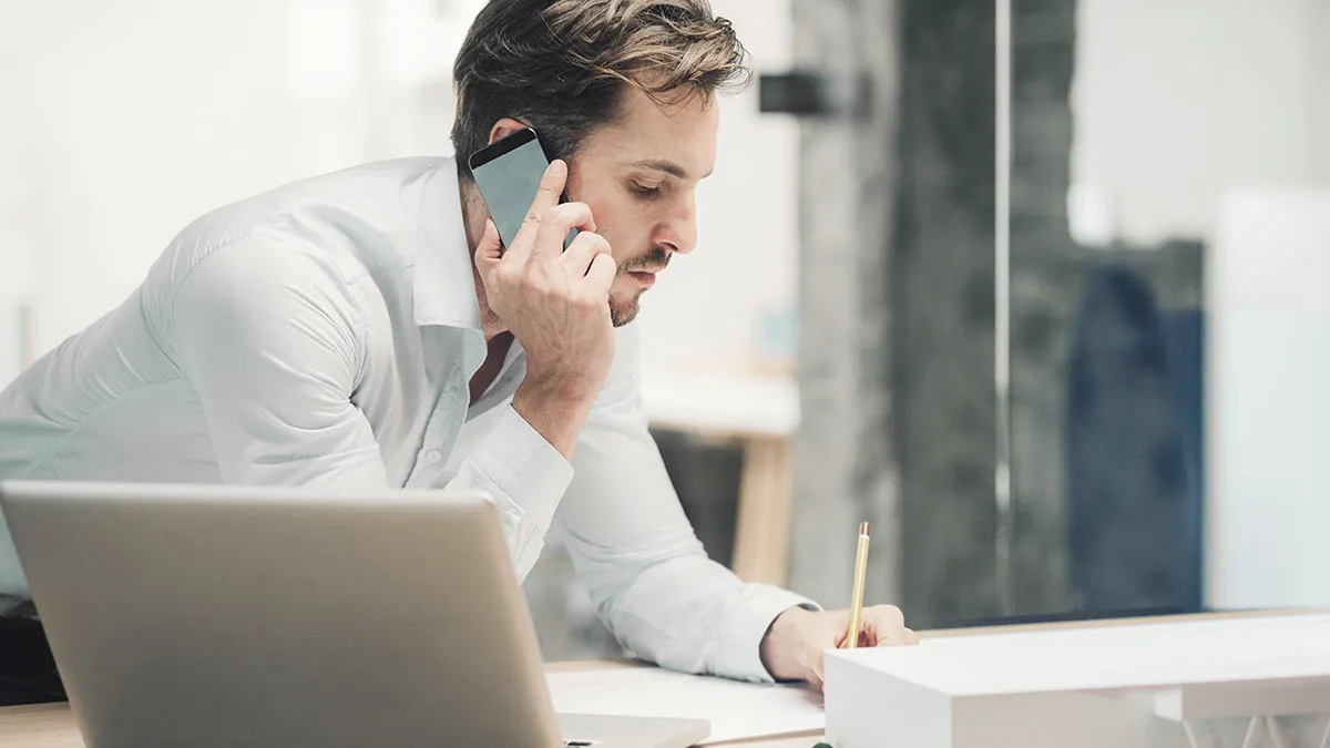 Ein Mann im weißen Hemd telefoniert am Schreibtisch mit dem Smartphone, während er Notizen macht, mit einem geöffneten Laptop im Vordergrund.