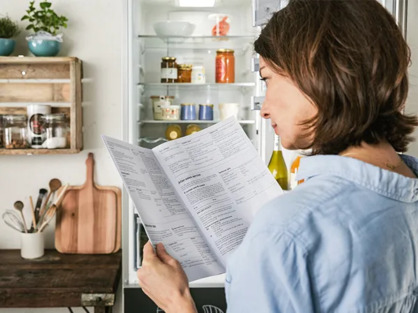Woman reading document in front of open refrigerator with kitchen items.