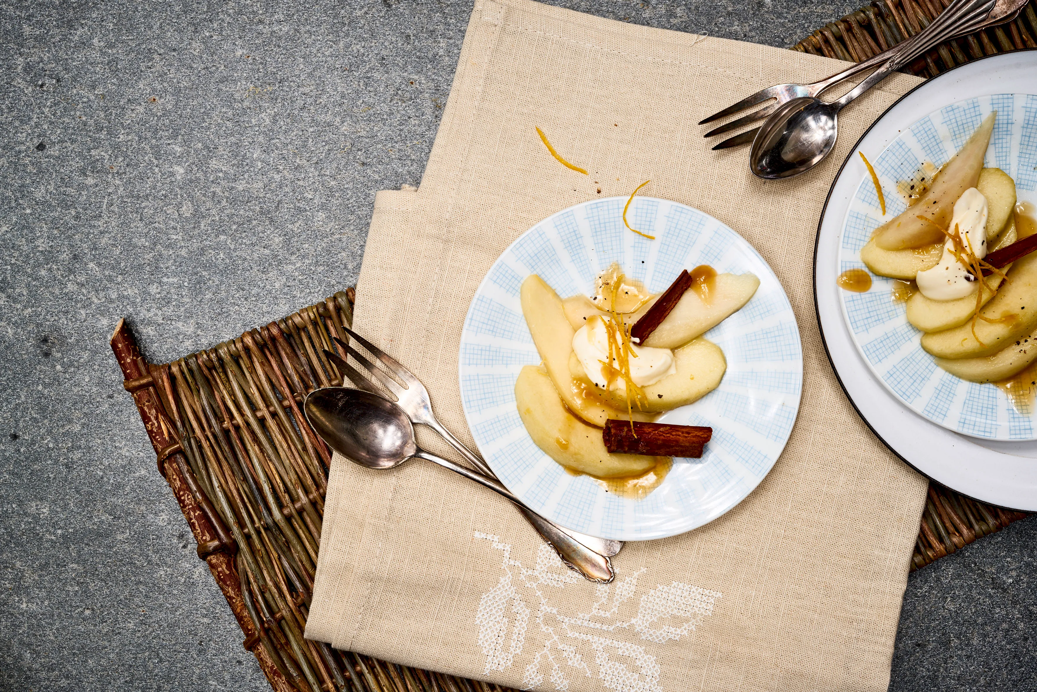 Piatti di pere cotte conditi con panna, bastoncini di cannella e scorza d'arancia, disposti su un tappetino intrecciato e un panno beige.