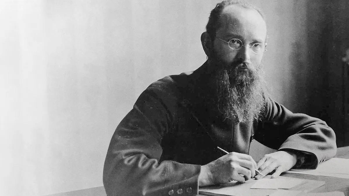 A man with a beard sits at a table, writing on a piece of paper in a black and white photograph.