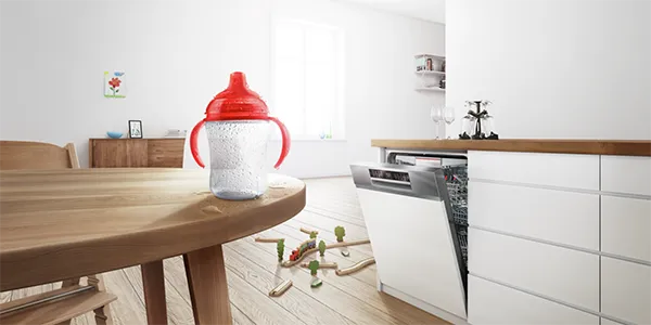 A baby bottle with a red top sits on a wooden table with water droplets, while a Bosch dishwasher is open in the background, and a toy train track is on the floor.