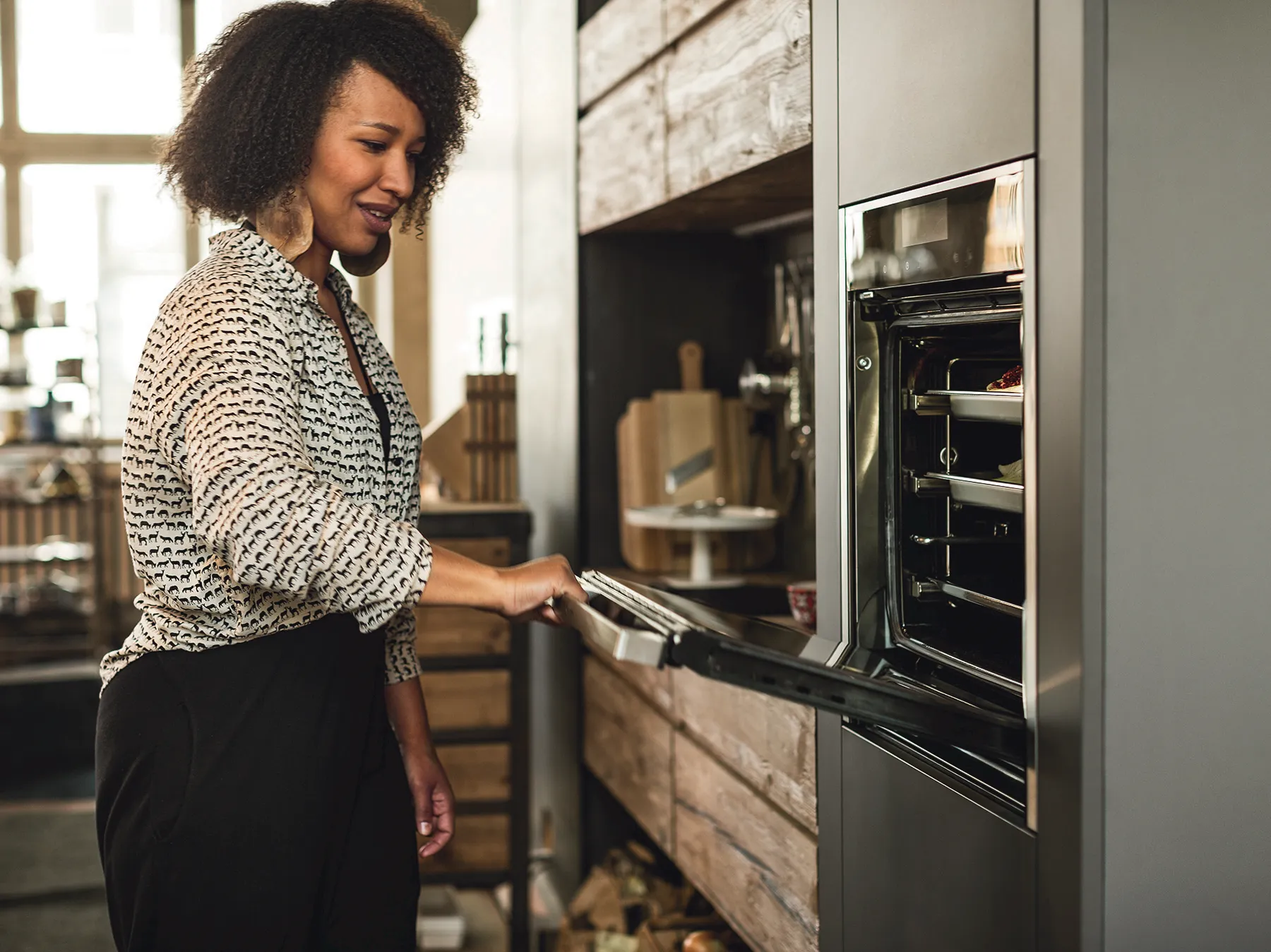 A smiling woman with dark, curly hair and large hoop earrings opens the door of a sleek, built-in oven in a rustic-modern kitchen. Inside the oven, trays hold food, including what appears to be a pie or casserole. The background features open wooden shelving with various kitchen items like cutting boards and a cake stand.