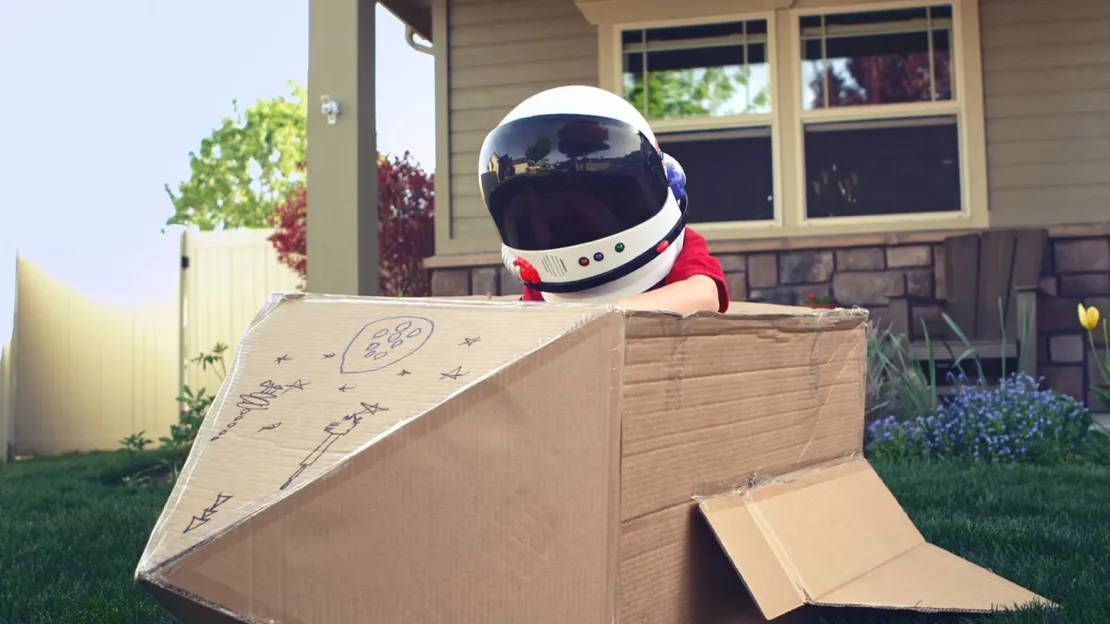Enfant portant un casque d'astronaute jouant dans un carton transformé en fusée dans un jardin devant une maison.