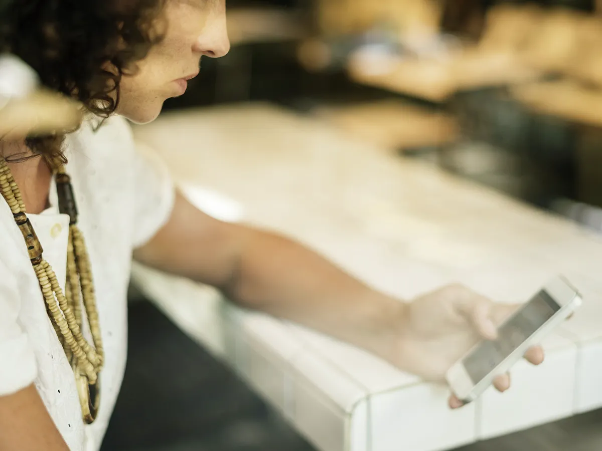 Person with curly hair and beaded necklace sitting at a table, looking at a smartphone held in their hand.