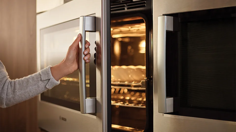 A person opens a Bosch stainless steel oven with a visible handle and warm interior lighting.