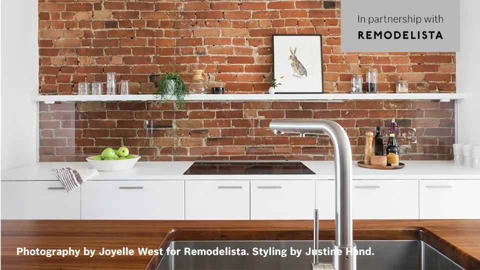 Modern kitchen with brick wall, white cabinets, and metal sink faucet; art on shelf.