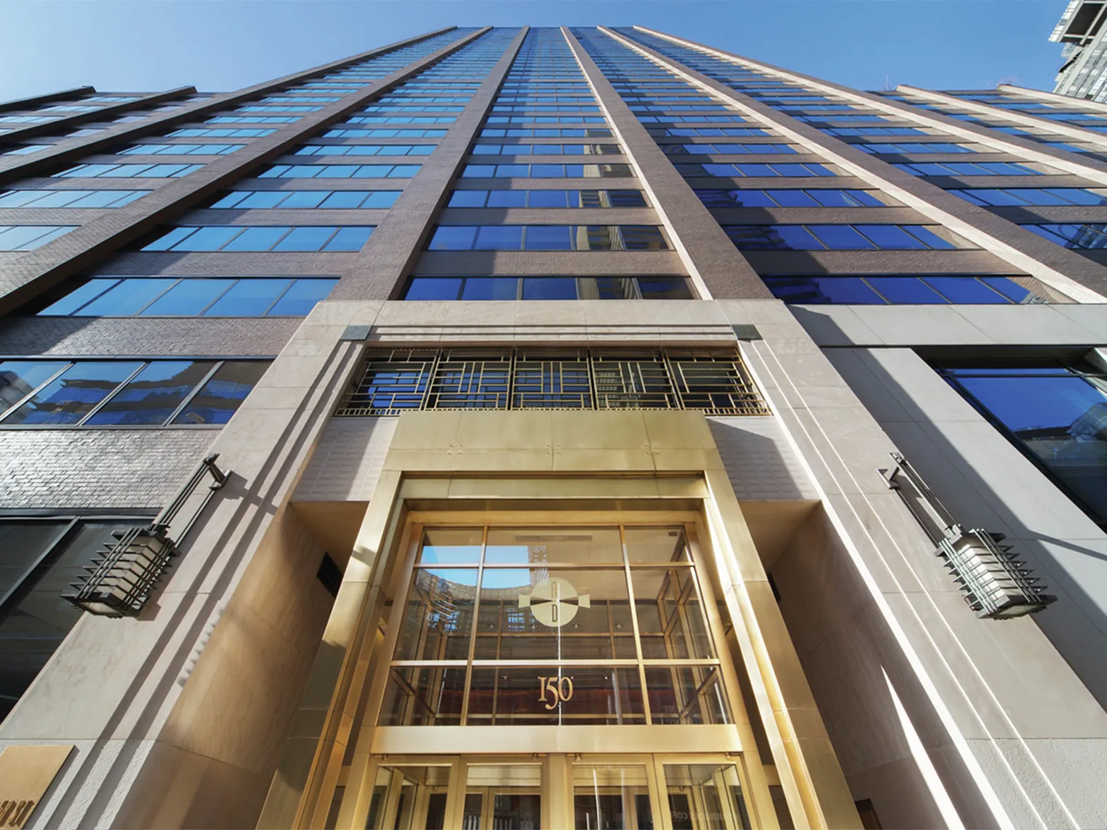 Tall glass skyscraper with a gold entrance, seen from below against a clear blue sky.