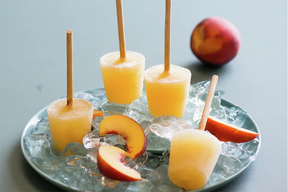 Frozen peach popsicles on a plate with ice cubes and a fresh peach.