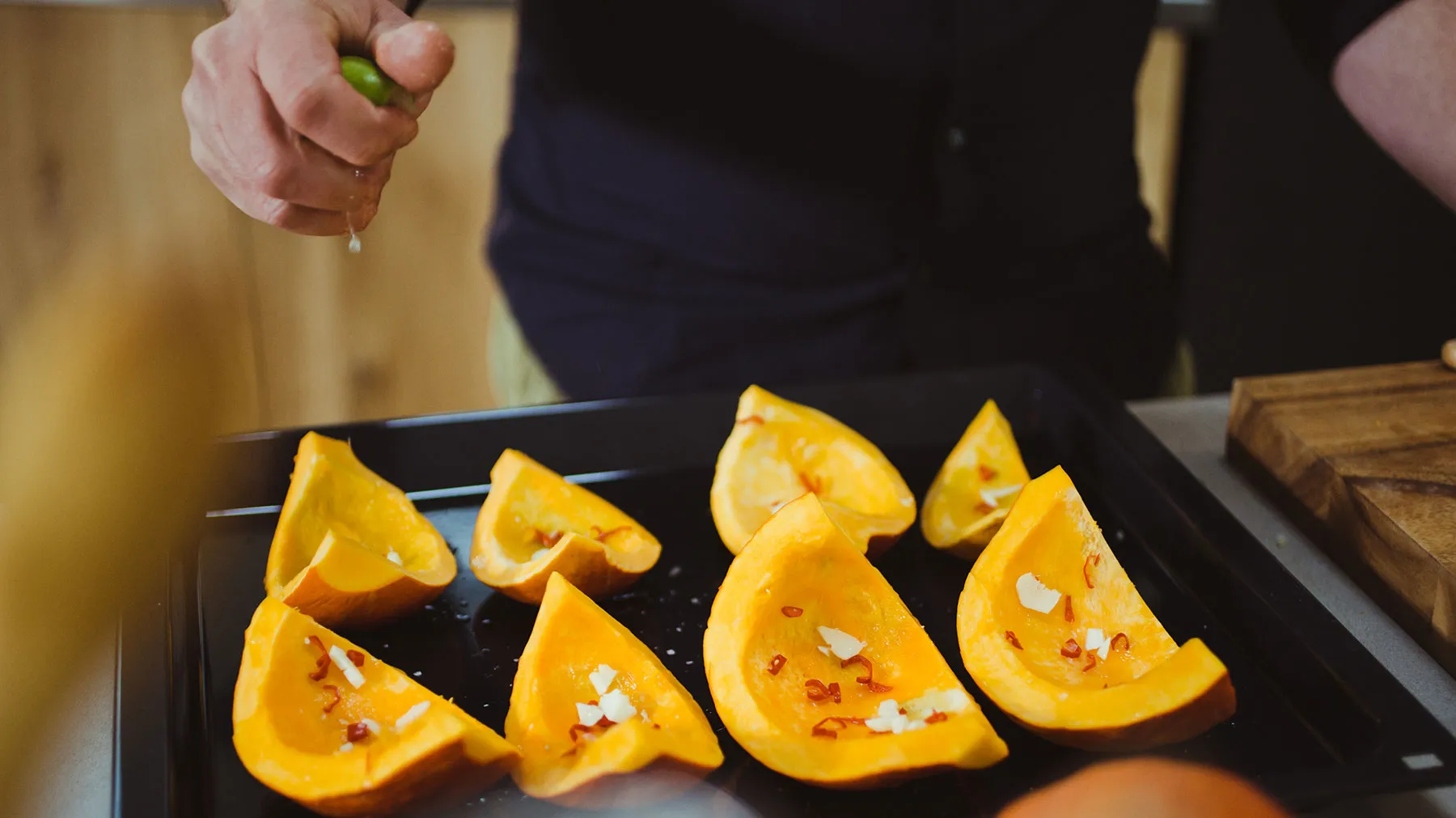 Hands cutting squash slices on a black tray, with a wooden cutting board in the background.