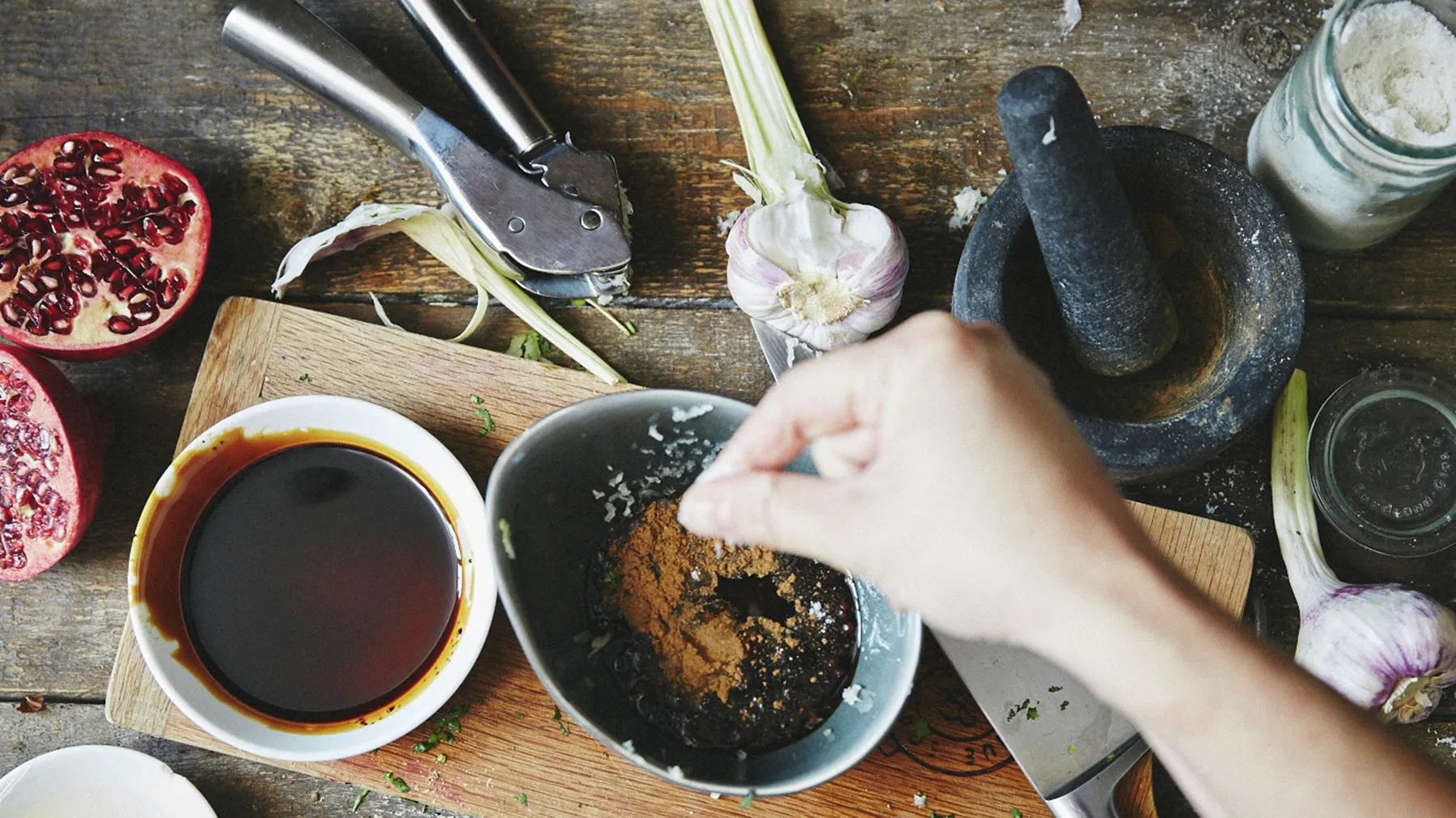 Cooking ingredients and tools on wooden table, including pomegranate, garlic, and a hand mixing something in a bowl.