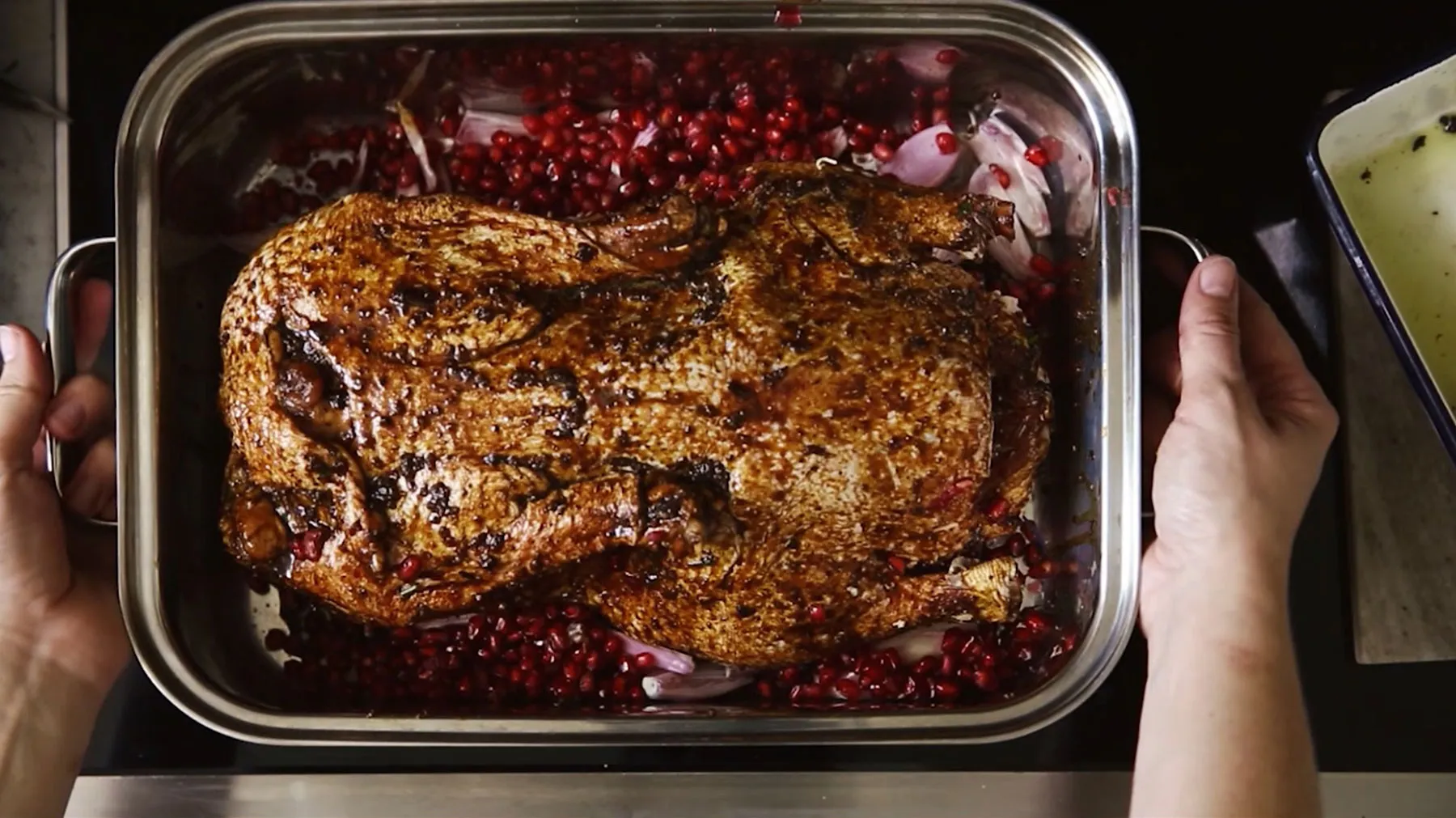 Roasted meat dish with pomegranate seeds in a baking tray, held by hands.