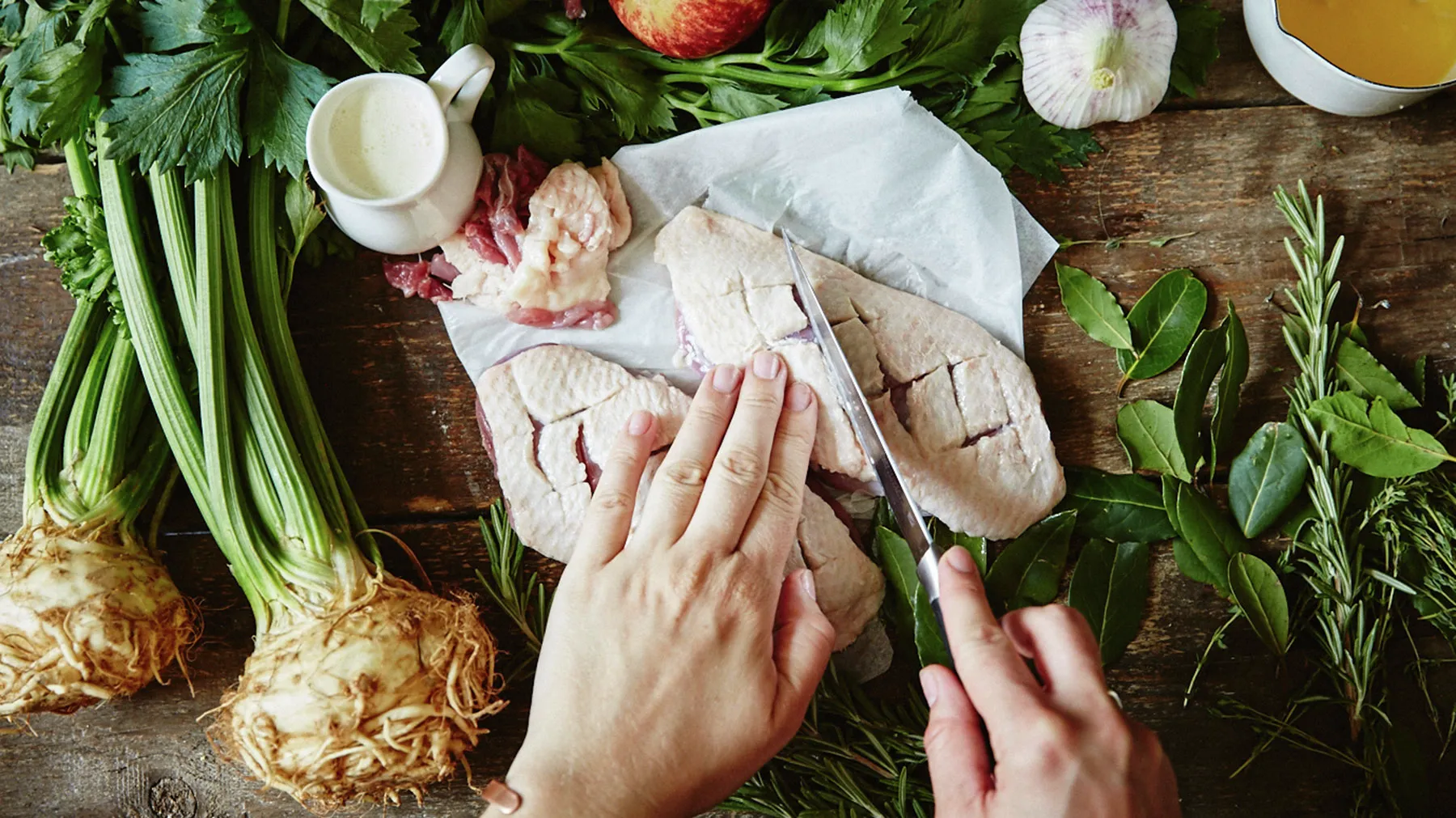 Hands preparing fresh ingredients - vegetables, herbs, and meat on a rustic wooden surface.