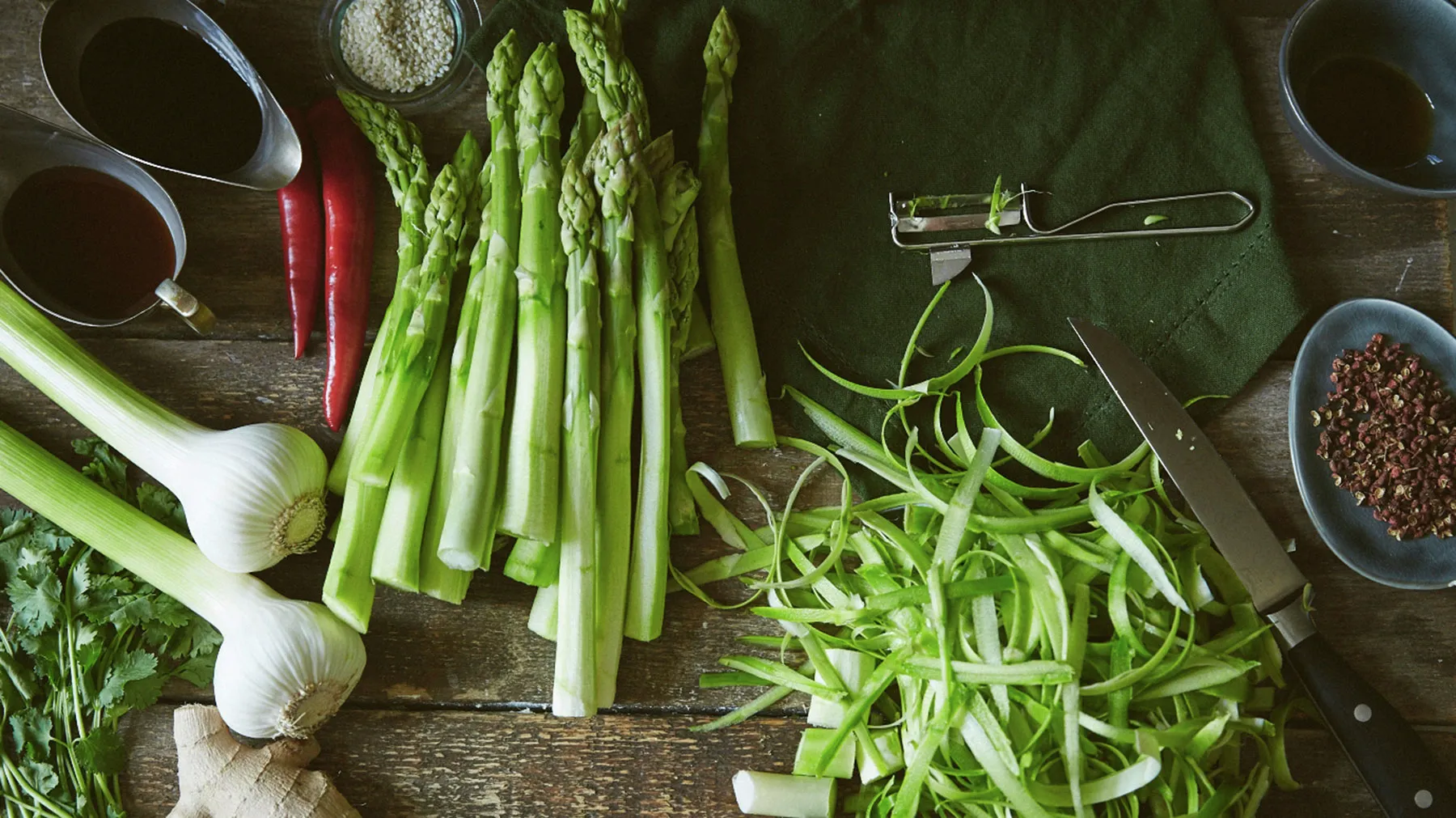 An overhead view of fresh green asparagus, red chilies, garlic, ginger, cilantro, and asparagus peelings arranged on a rustic wooden table, alongside a peeler, knife, and bowls of sauces and spices.