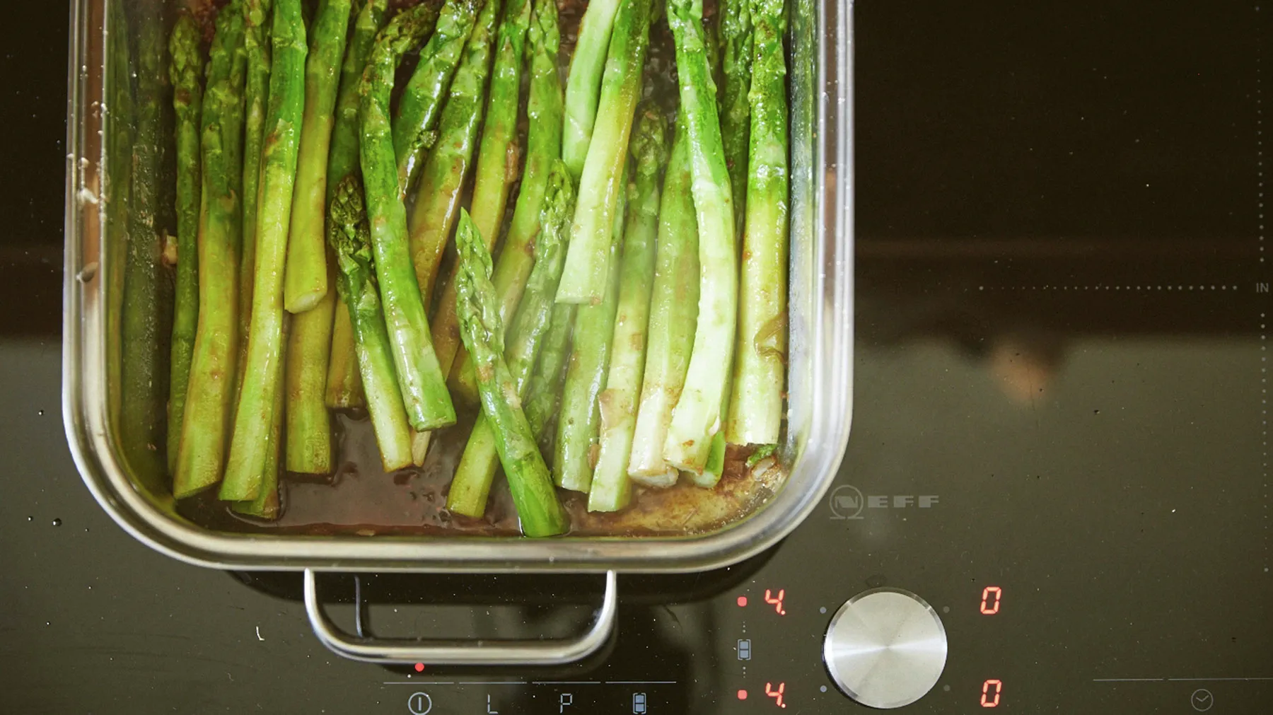 A rectangular stainless steel pan filled with green asparagus spears in a dark sauce is cooking on a black Nespresso induction cooktop with red digital controls.