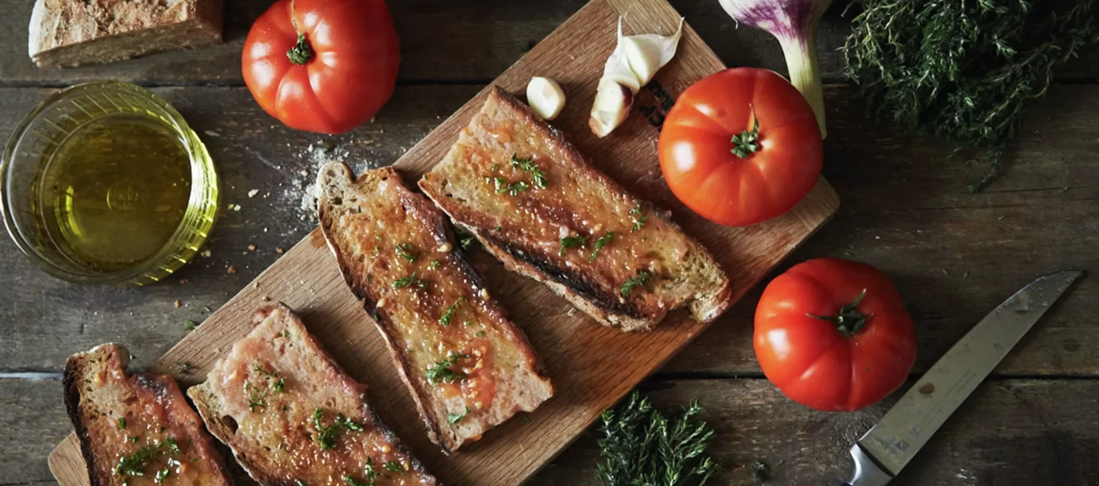 Sliced bread, tomatoes, garlic, olive oil, and herbs on a wooden cutting board.