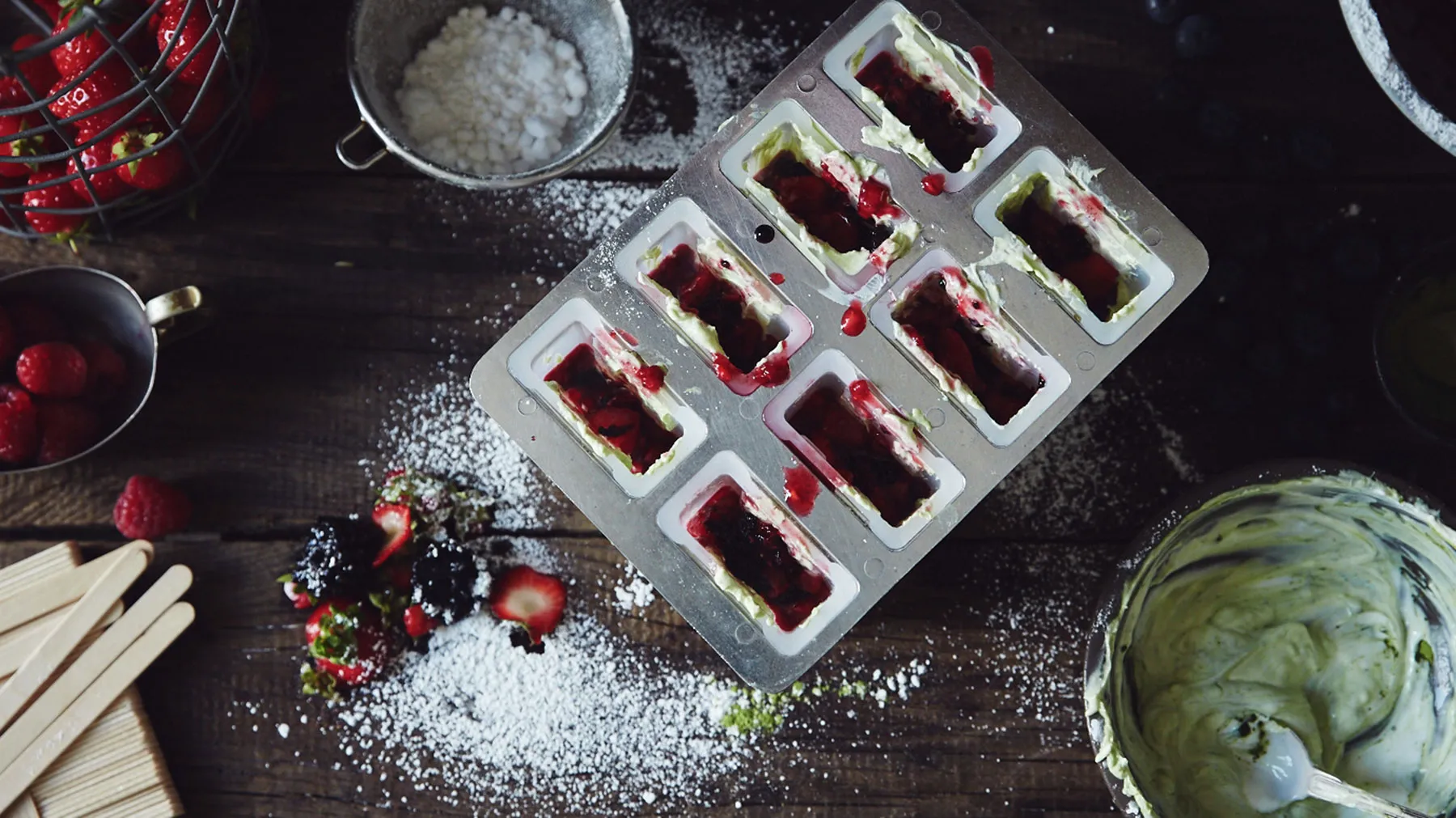 An overhead view of a metal popsicle mold filled with green and red fruit mixture, surrounded by fresh berries, powdered sugar, and wooden popsicle sticks on a rustic wooden table.