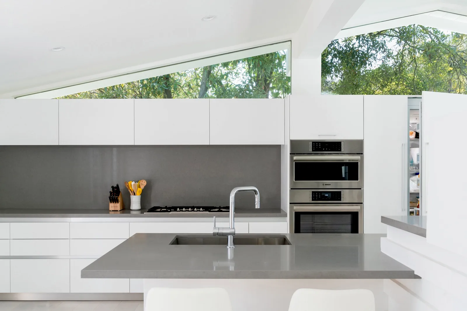 Modern kitchen with gray countertop, Bosch built-in appliances, and large windows with greenery outside.