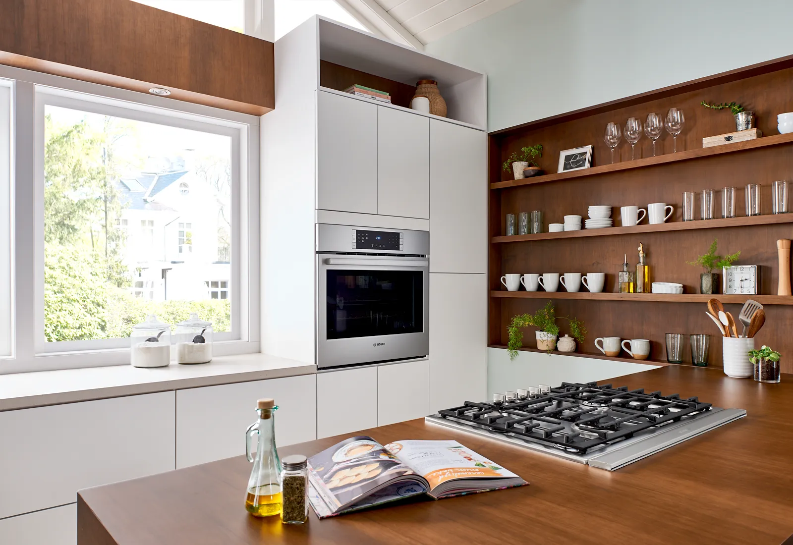Modern kitchen with wooden shelves, Bosch gas stove, Bosch wall oven, and open window overlooking greenery.