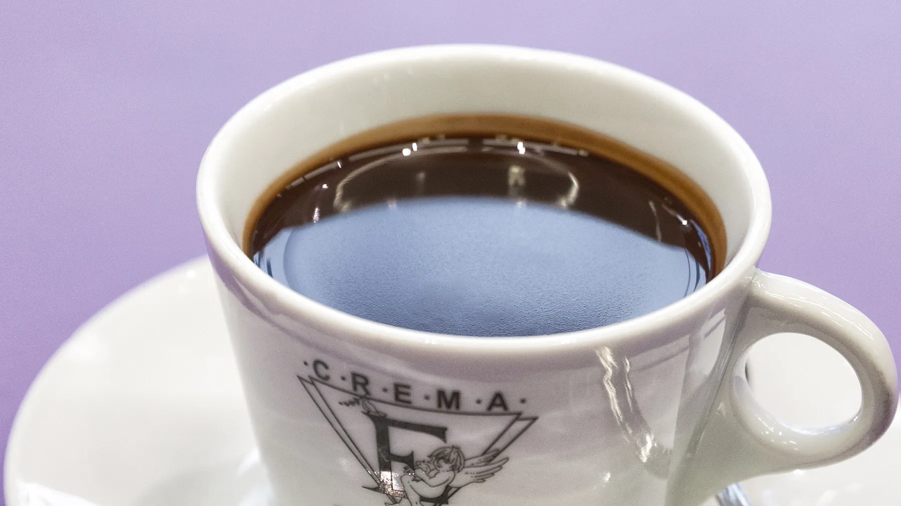 Weiße Keramik Tasse mit Crema Logo, gefüllt mit dunklem Kaffee, vor einem lila Hintergrund.