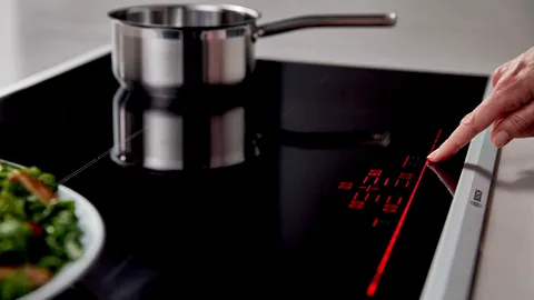 Stainless steel pot on Bosch induction cooktop, fresh salad in foreground, hand adjusting temperature.