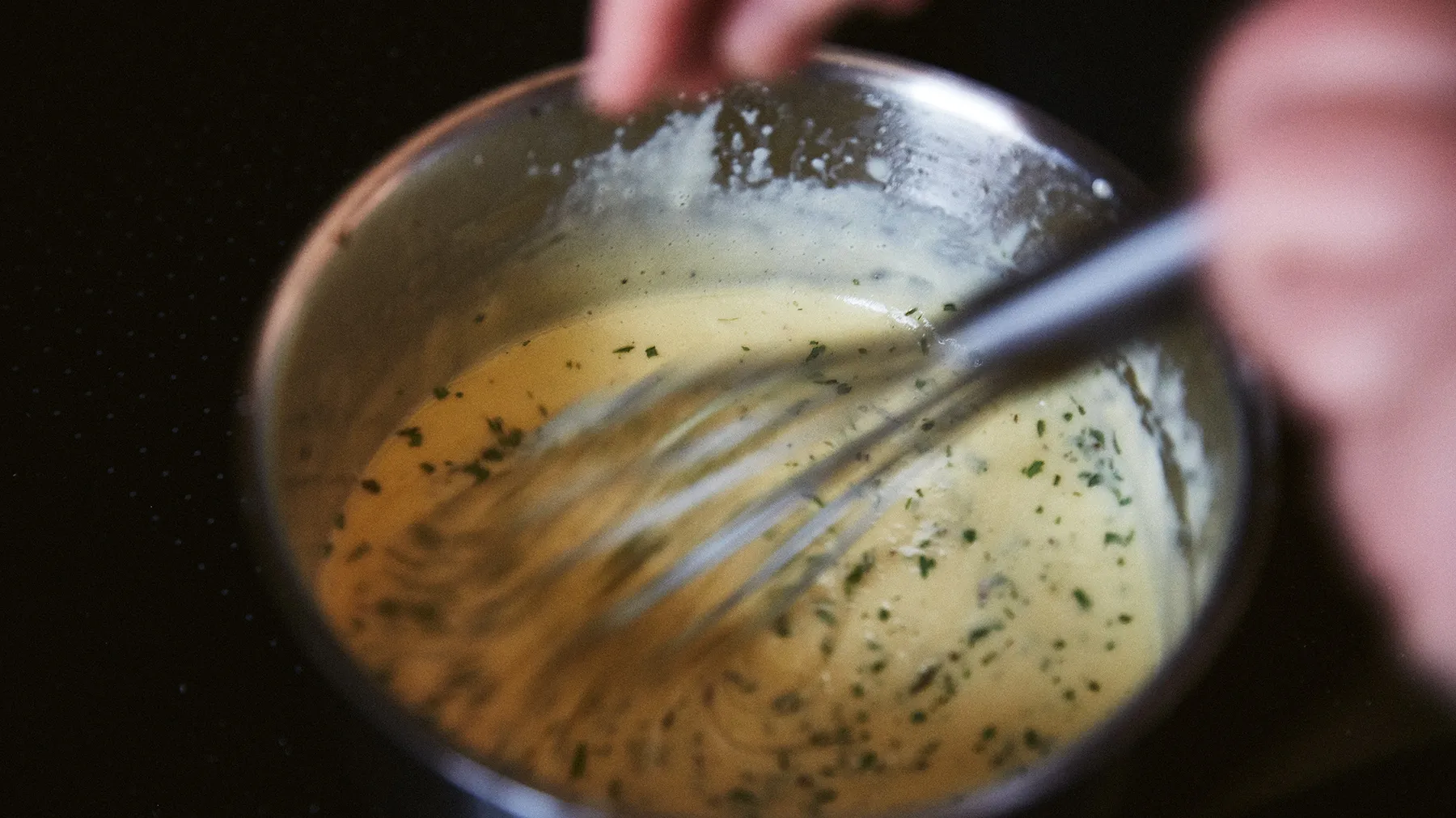 Mani che fischiano un liquido ghiacciato e dorato in una ciotola di vetro su uno sfondo scuro.