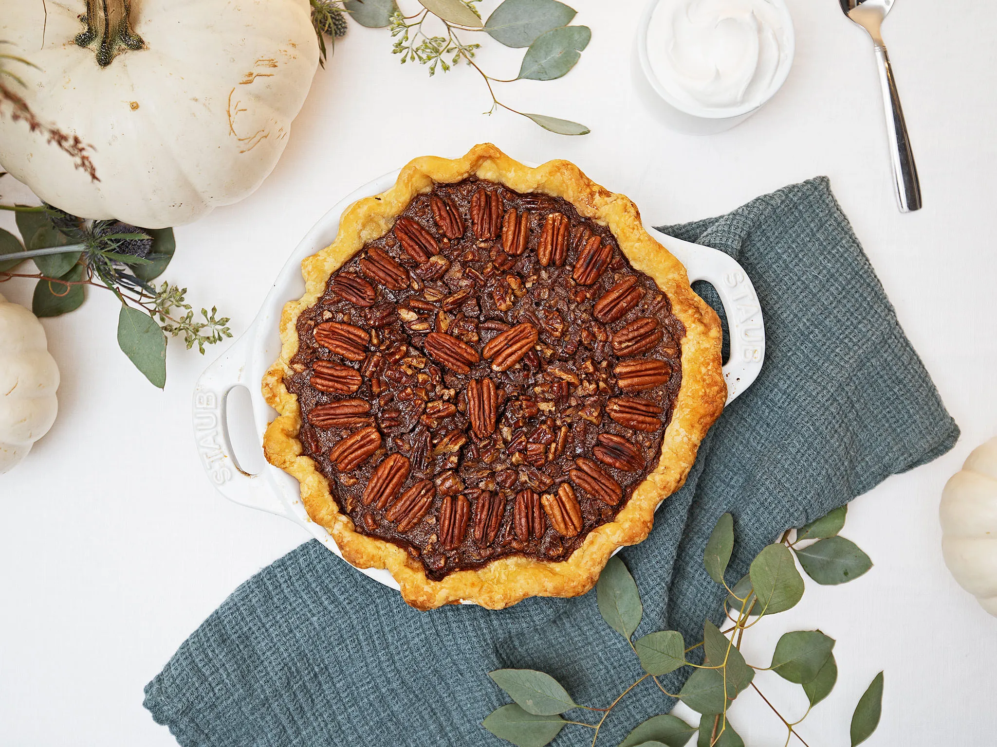 Pecan pie with flaky crust, surrounded by greenery and kitchen towel on white background.