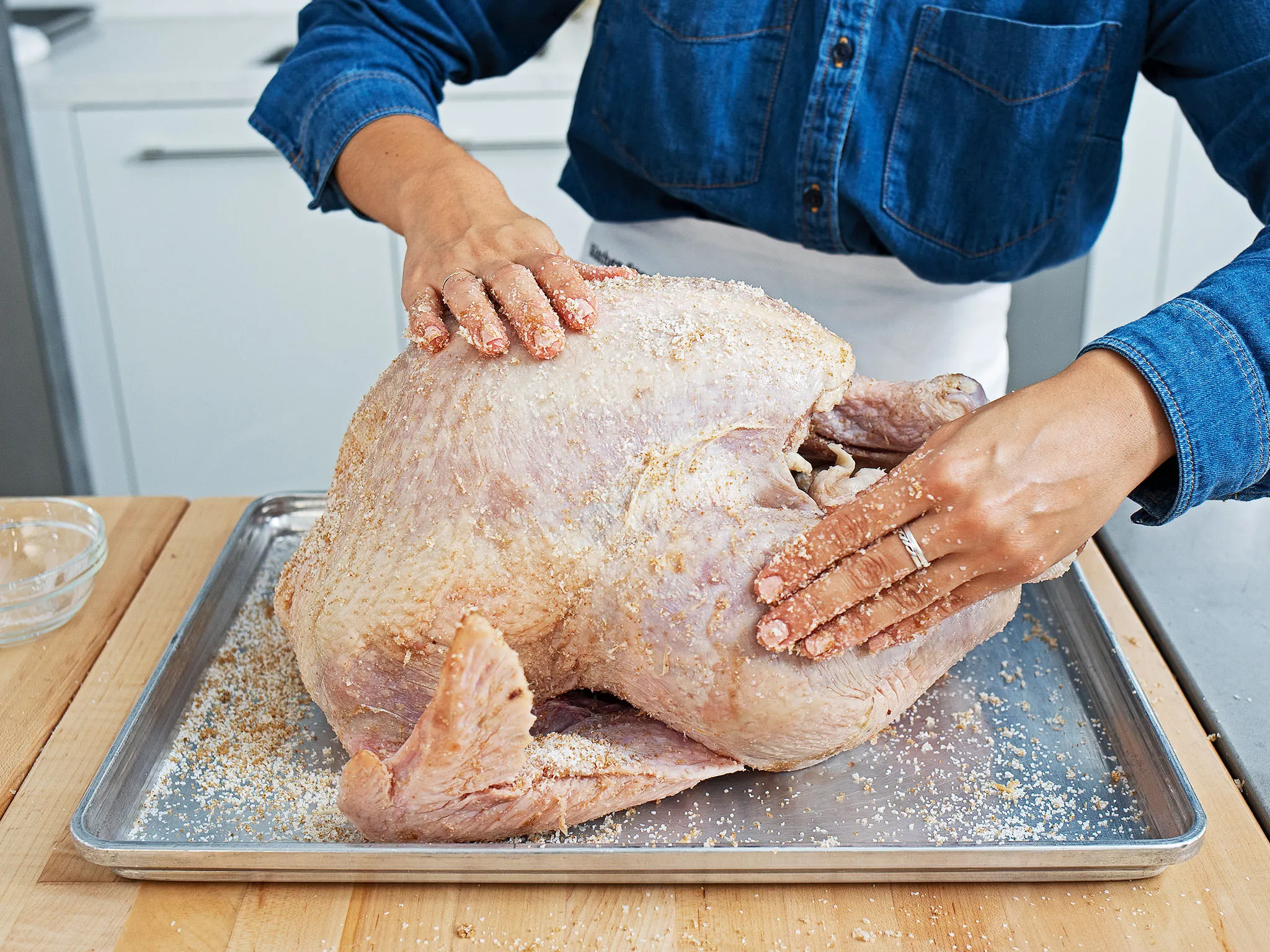 A person seasoning a raw turkey on a metal tray in a kitchen setting.