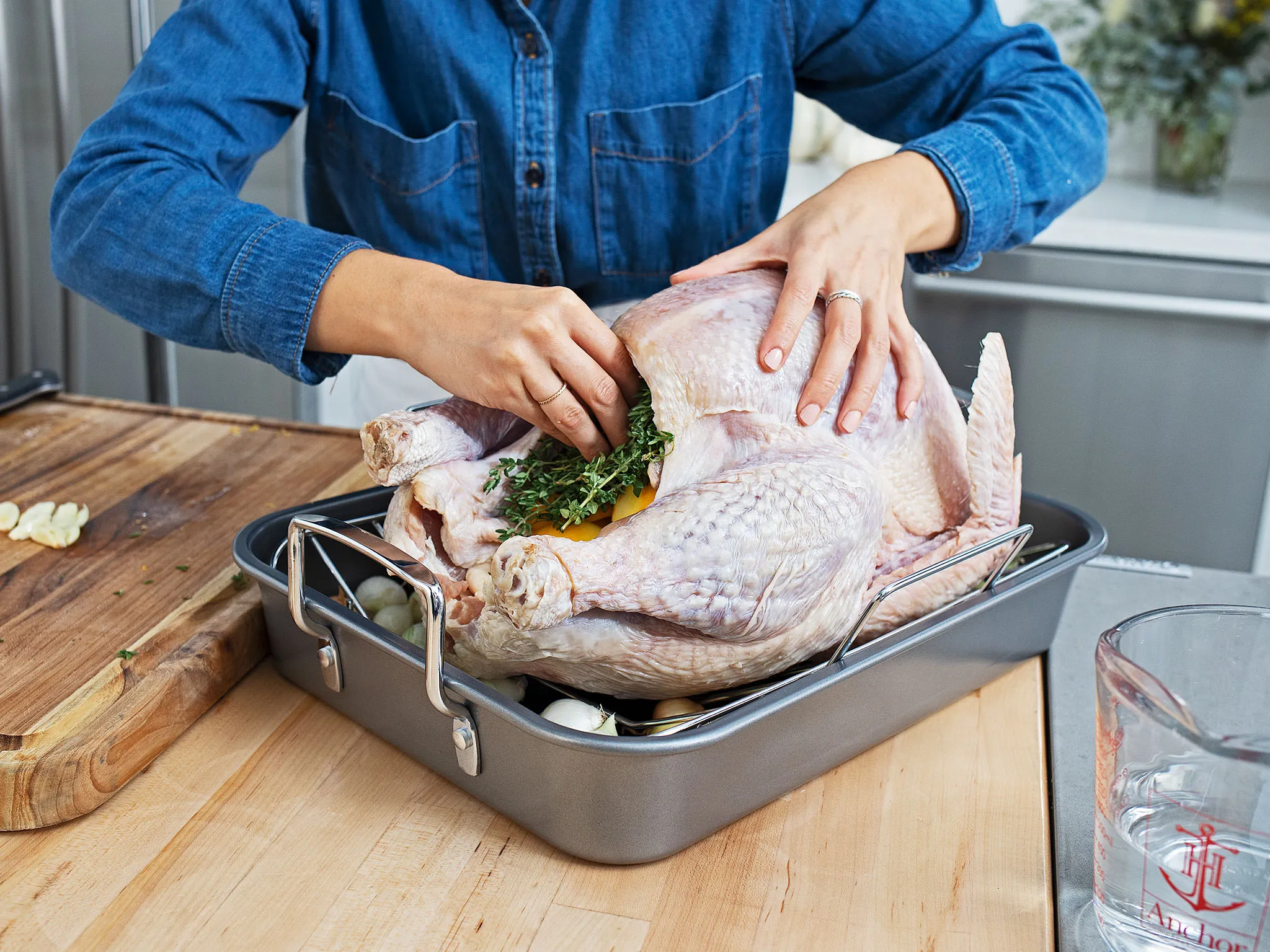 Person stuffing a turkey with herbs in a metal roasting pan on a wooden counter.