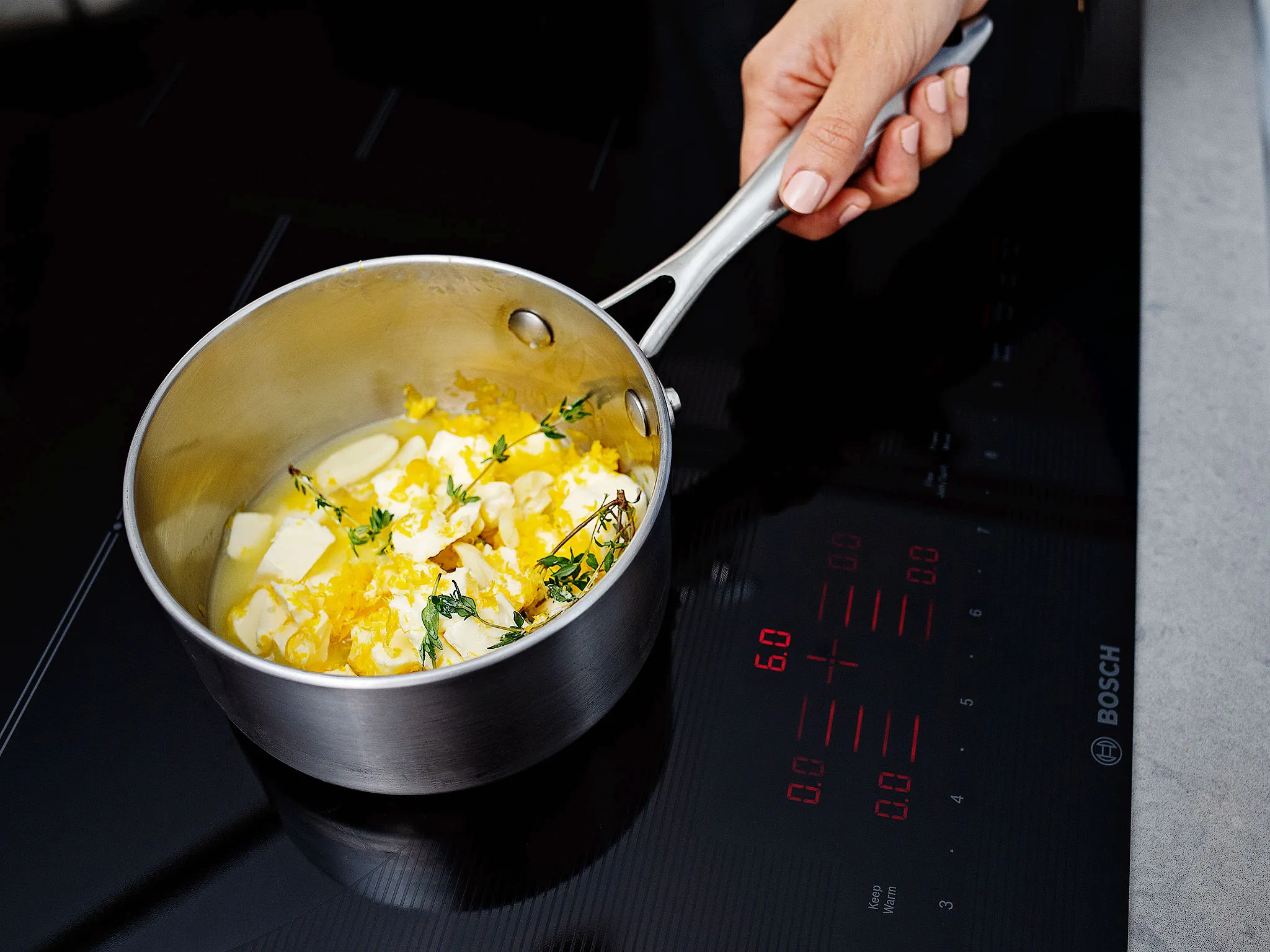 Cooking pot on Bosch induction cooktop with butter and herbs, hand adjusting the pot.