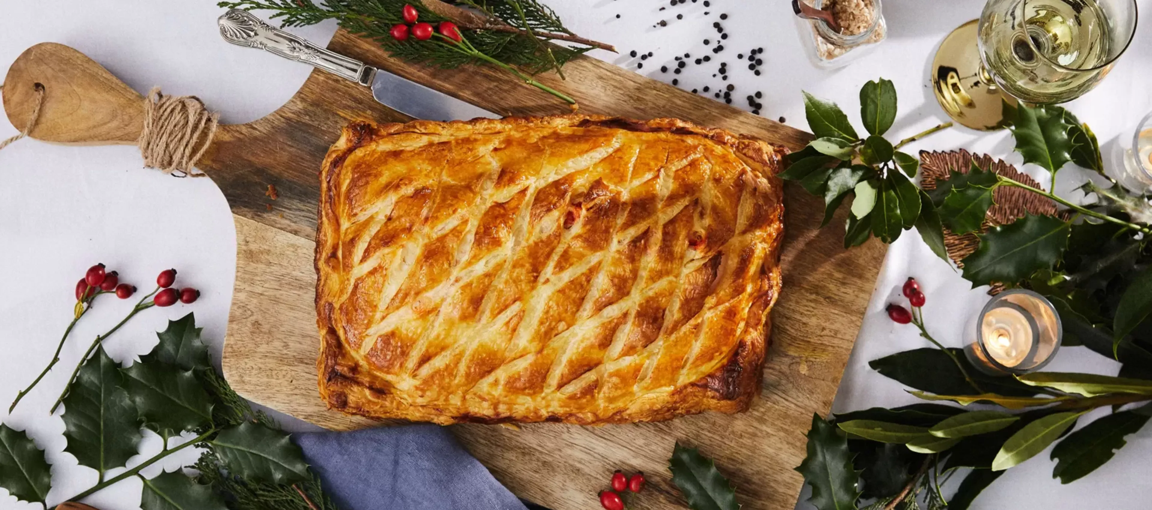 Festive holiday meal with baked pastry, herbs, spices, and wine glasses on a wooden board.