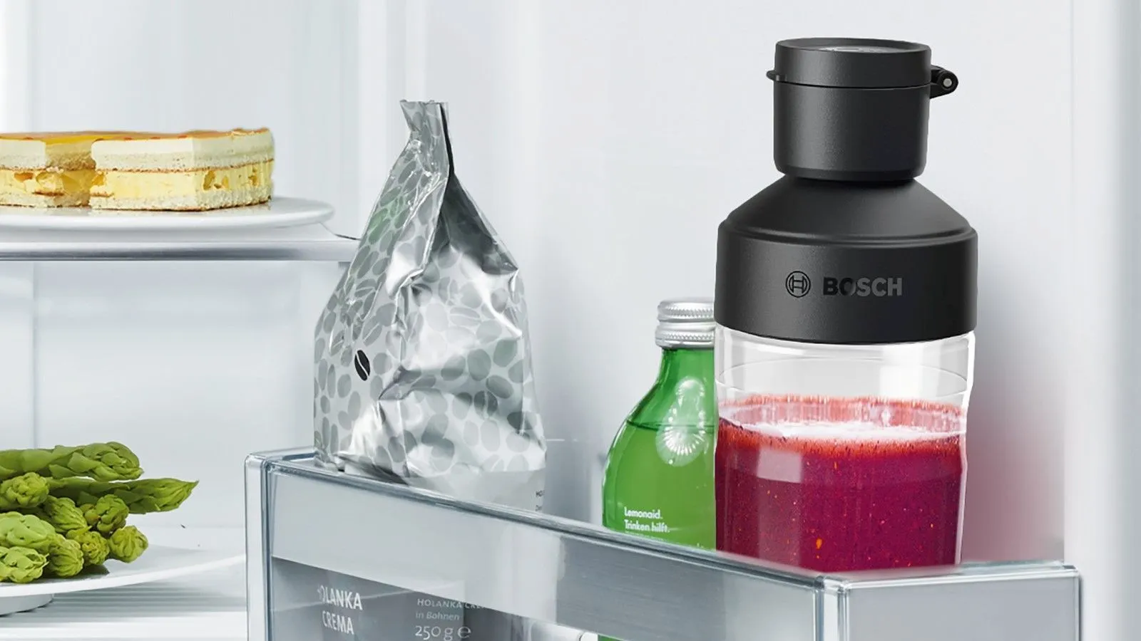 Interiér chladničky s tortou, zeleninou a mixérom Bosch s červeným smoothie.